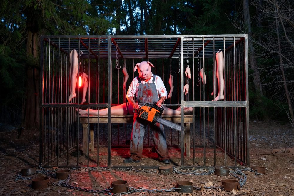 A butcher in a pig mask and bloodied apron standing in a steel cage hung with meat hooks